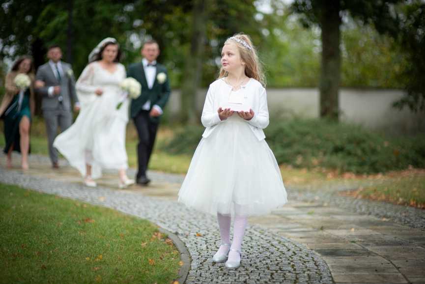Dziewczynka w białej sukni prowadzi młodą parę do Kościoła